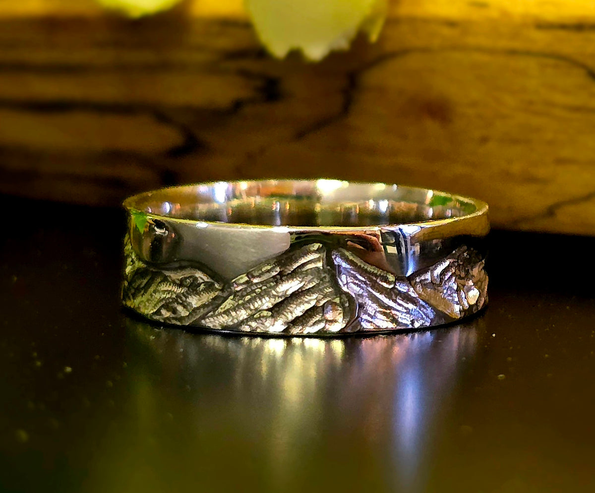 A polished silver ring with a textured mountain landscape engraving, resting on a dark reflective surface with a warm wooden background.