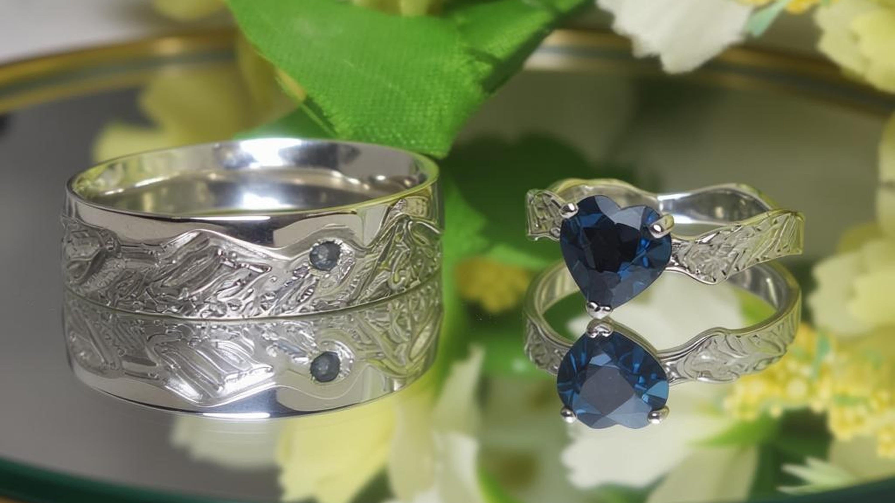 Two silver rings with blue gemstones on a reflective surface with flowers in the background
