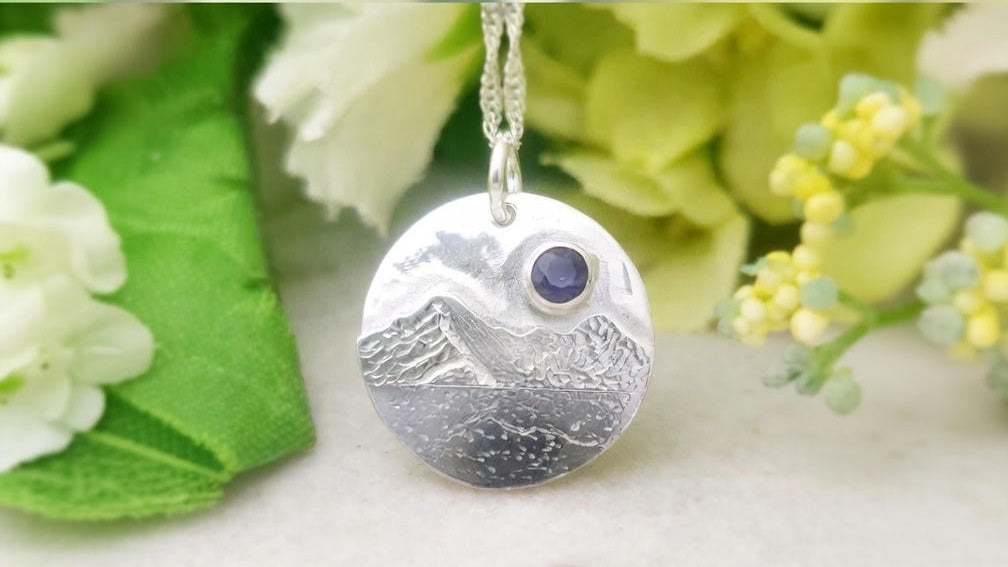 Silver mountain pendant necklace with a blue gemstone on a marble surface with flowers in the background